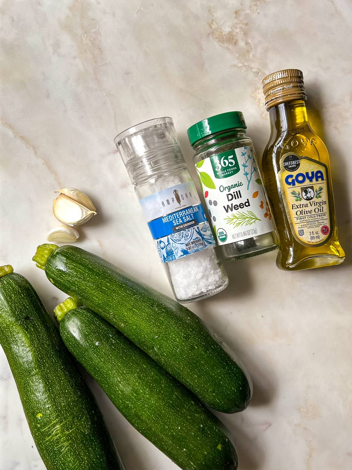 Overhead view of zucchini, garlic cloves, olive oil, salt, and dried dill laid out on a white surface.