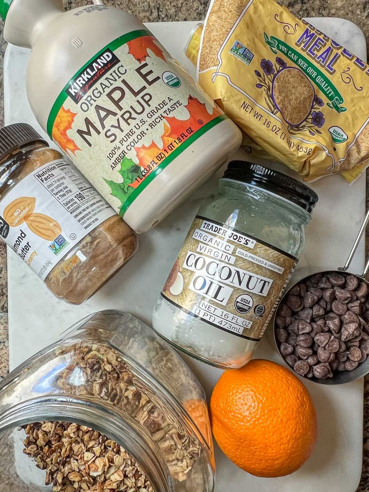 Ingredients for homemade granola bars without peanut butter, including almond butter, coconut oil, maple syrup, chocolate chips, granola, ground flaxseed, and an orange, all arranged on a countertop.
